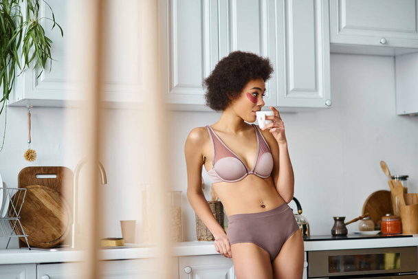 femme afro-américaine bouclée avec des patchs debout en sous-vêtements et boire du café dans une tasse - Photo, image