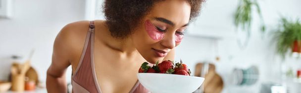 joven afroamericana mujer en lencería oliendo fresas frescas en la cocina, pancarta - Foto, Imagen