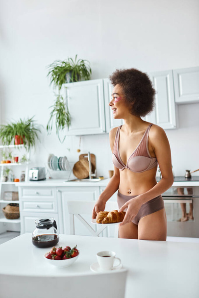 heureuse femme afro-américaine en lingerie servant croissants et fraises pour le petit déjeuner - Photo, image