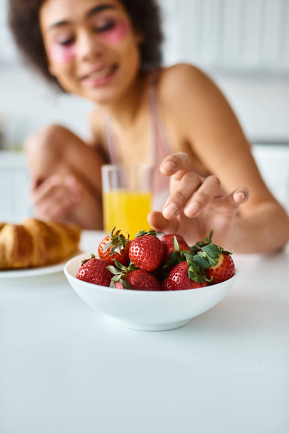 θολή Αφροαμερικανή γυναίκα φθάνει φρέσκες φράουλες κατά τη διάρκεια του πρωινού στην κουζίνα - Φωτογραφία, εικόνα