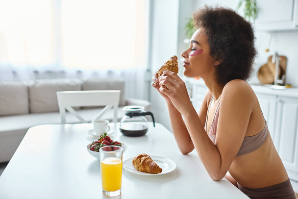 sorrindo Africano americano mulher segurando croissant recém-assado durante o café da manhã na cozinha - Foto, Imagem