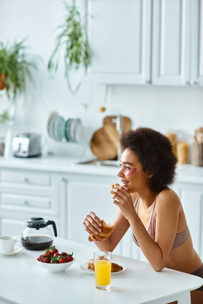 キッチンで朝食中に新鮮なクロワッサンを抱えている陽気なアフリカ系アメリカ人女性 - 写真・画像