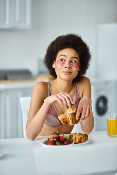 pensive african american woman holding freshly baked croissant during breakfast in kitchen - Photo, Image