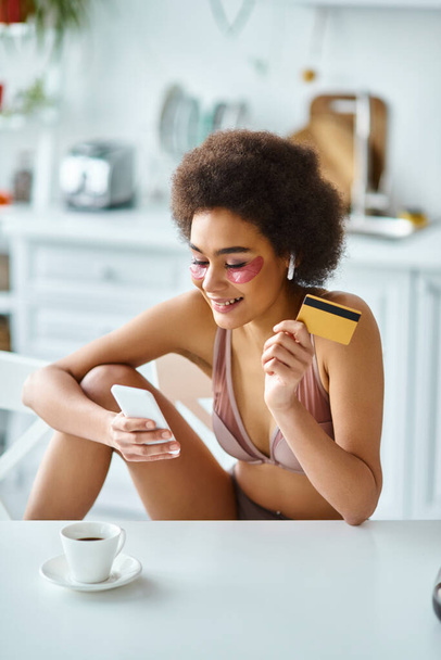 Sonriente mujer afroamericana de compras en línea con tarjeta de crédito y teléfono inteligente en la cocina - Foto, imagen