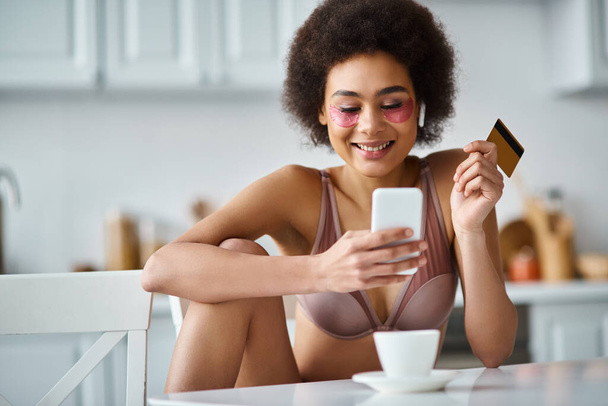 ポジティブなアフリカ系アメリカ人女性がキッチンでクレジットカードとスマートフォンでオンラインショッピング - 写真・画像