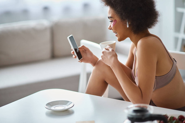 Усміхнена афроамериканка у спідній білизні з плямами очей насолоджується кавою та переглядом смартфона - Фото, зображення