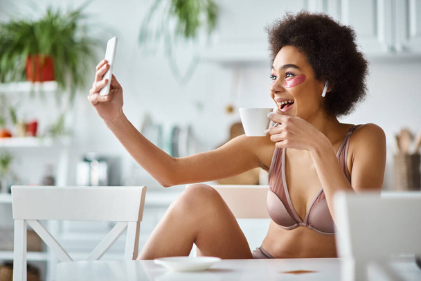 Mujer afroamericana sonriente en lencería con parches para los ojos tomando selfie con taza de café - Foto, Imagen