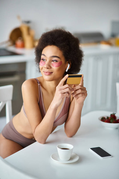 uśmiechnięta Afroamerykanka w słuchawkach z łatami pod oczami robiąca zakupy kartą kredytową - Zdjęcie, obraz