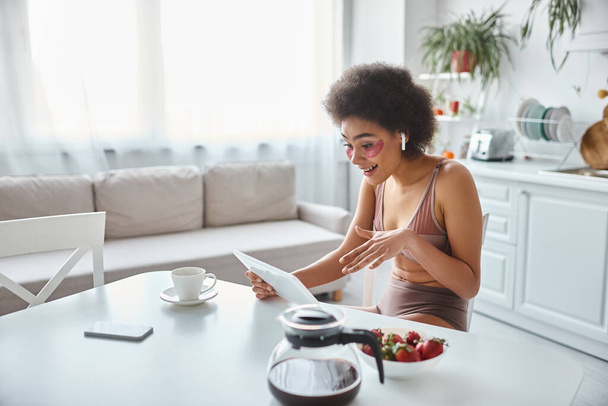 sorridente mulher afro-americana em lingerie e olhos remendos ter vídeo chat na cozinha moderna - Foto, Imagem