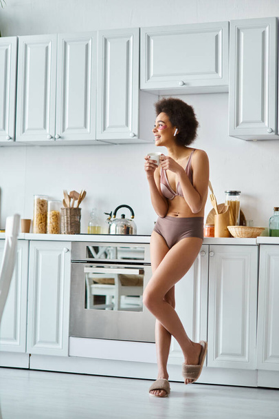 Moment serein de femme afro-américaine heureuse dans les écouteurs et la lingerie sirotant le café du matin - Photo, image