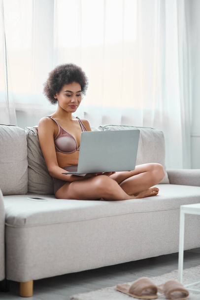молодой африканский американец в нижнем белье работает удаленно на ноутбуке, сидя уютный диван дома - Фото, изображение