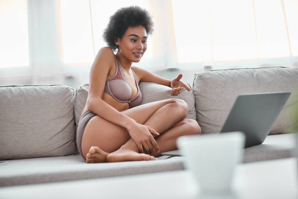 africká americká žena ve spodním prádle sedí na gauči a usmívá se při pohledu na laptop, víkend - Fotografie, Obrázek