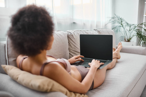 вид сзади на африканца в нижнем белье, который скромно работает на ноутбуке, сидя дома на уютном диване - Фото, изображение