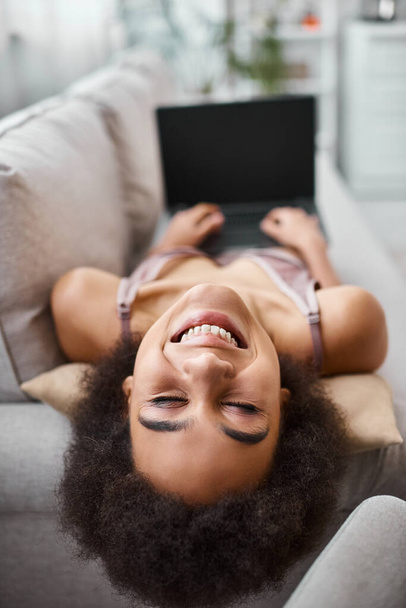 позитивний афроамериканський американець у спідній білизні працює дистанційно на ноутбуці, сидячи затишним диваном вдома - Фото, зображення