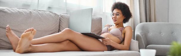 баннер африканской американки в нижнем белье и очках, работающих дистанционно на ноутбуке в гостиной - Фото, изображение