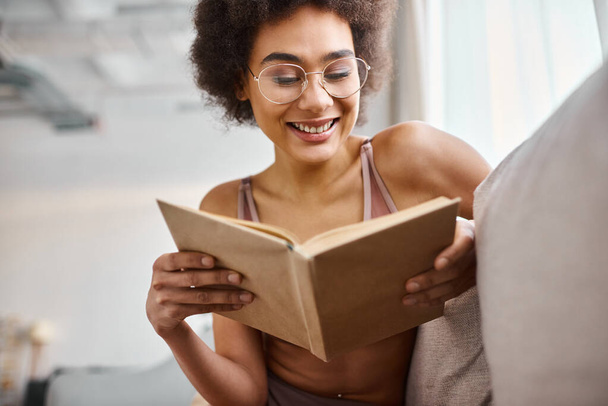 veselé a kudrnaté africké Američanky čtení knihy ve spodním prádle na pohodlné pohovce - Fotografie, Obrázek