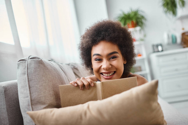 весела і молода афроамериканка тримає книгу і лежить на дивані вдома, насолоджуйтесь - Фото, зображення