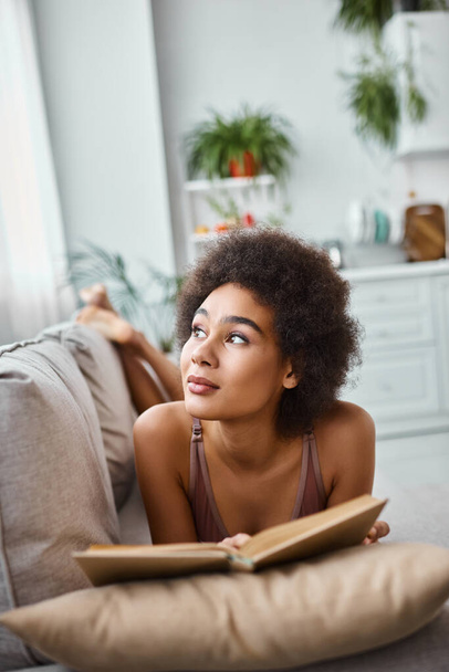 ονειρεμένη και νεαρή Αφροαμερικανή που διαβάζει ένα βιβλίο με εσώρουχα και ξαπλώνει σε έναν άνετο καναπέ. - Φωτογραφία, εικόνα