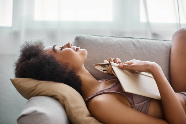 vista lateral de la ropa interior de mujer afroamericana joven y complacida acostada con libro abierto en un cómodo sofá - Foto, Imagen