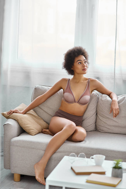 dreamy young african american woman with curly hair lounging in lingerie on a sofa at home - Photo, Image