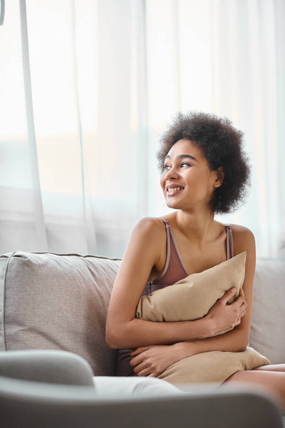 mladá africká americká žena s kudrnatými vlasy relaxaci na gauči ve spodním prádle, usmívá se a dívá se jinam - Fotografie, Obrázek