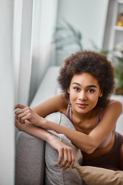 jolie femme américaine africaine avec les cheveux bouclés relaxant sur le canapé en lingerie, regardant la caméra - Photo, image
