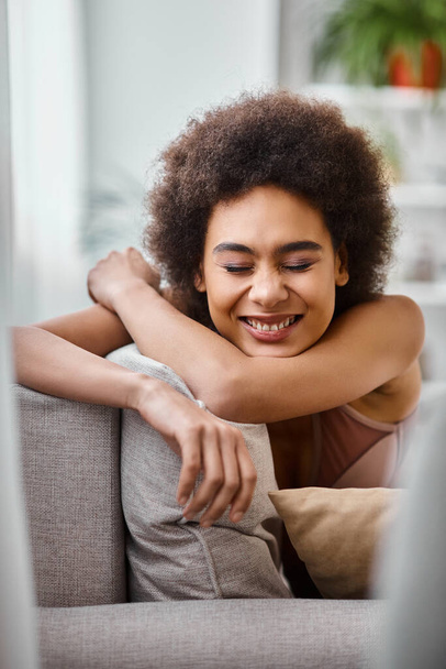 onnellinen afrikkalainen amerikkalainen nainen, jolla on kiharat hiukset rentouttava sohvalla alusvaatteita, hymyillen suljetuin silmin - Valokuva, kuva