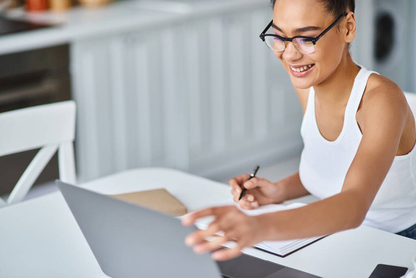 boldog afro-amerikai nő szemüvegben dolgozik otthonról távolról a laptopján, jegyzetel - Fotó, kép