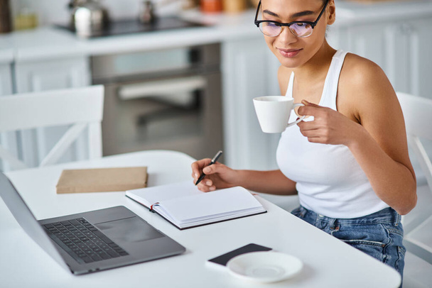 lockige Afroamerikanerin mit Brille, die von zu Hause aus ferngesteuert an ihrem Laptop arbeitet und Kaffee schlürft - Foto, Bild