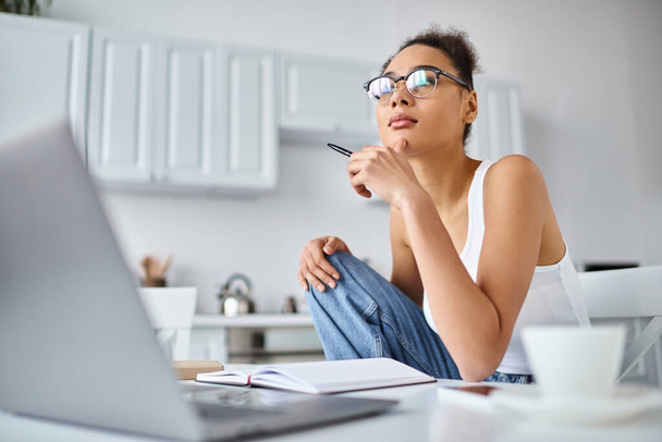 pensive african american woman in glasses working from home remotely on her laptop, cup of coffee - Photo, Image