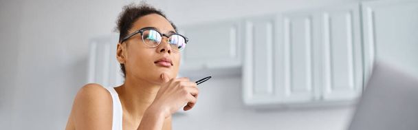 Femme afro-américaine coûteuse dans des lunettes travaillant à distance de la maison et tenant un stylo, une bannière - Photo, image