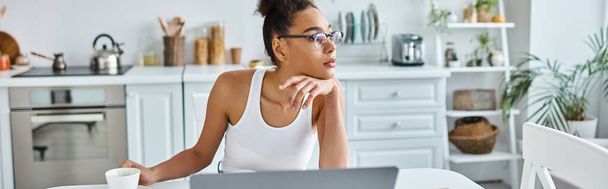 Pensive αφροαμερικανή γυναίκα σε γυαλιά κάθεται στο γραφείο με φορητό υπολογιστή και φλιτζάνι καφέ, πανό - Φωτογραφία, εικόνα