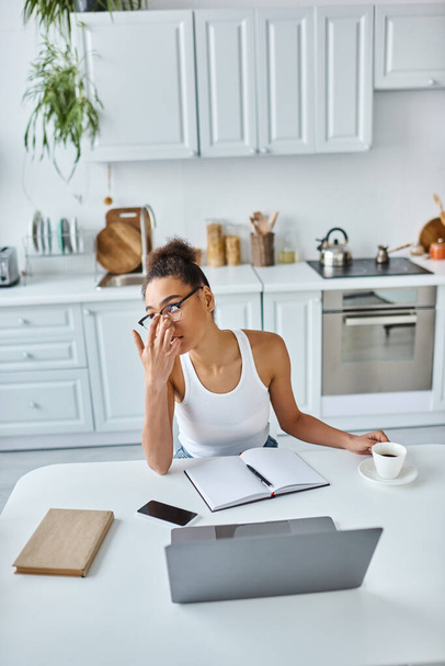 pensive african american woman adjusting glasses and sitting at desk with laptop and cup of coffee - Photo, Image
