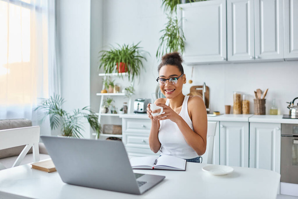 femme afro-américaine positive dans des lunettes tenant une tasse de café près d'un ordinateur portable sur le bureau, travail à distance - Photo, image
