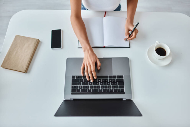 bovenaanzicht van Afrikaanse Amerikaanse vrouw met pen in de hand met behulp van laptop naast kopje koffie op het bureau - Foto, afbeelding