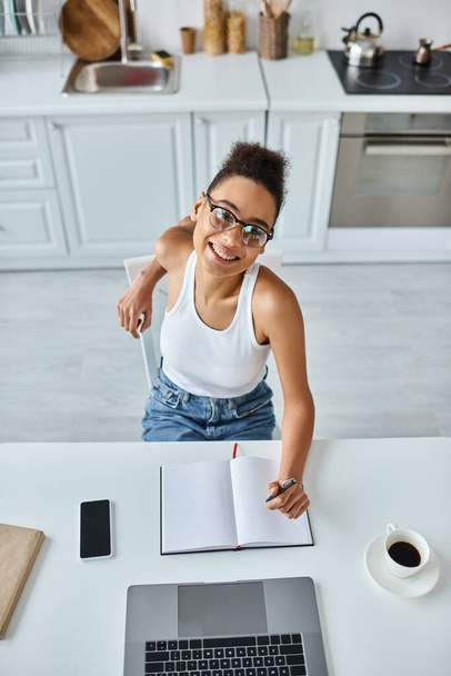 vue du haut de femme afro-américaine heureuse avec stylo à la main en utilisant un ordinateur portable à côté de la tasse de café sur le bureau - Photo, image