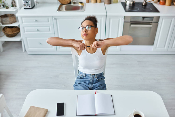 vue de dessus de femme afro-américaine fatiguée avec stylo assis au bureau avec tasse de café et ordinateur portable - Photo, image