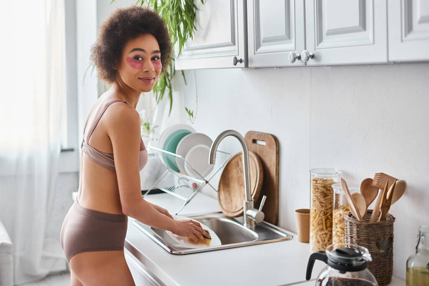 femme afro-américaine gaie en lingerie avec des patchs pour les yeux plaque de lavage avec éponge dans la cuisine - Photo, image