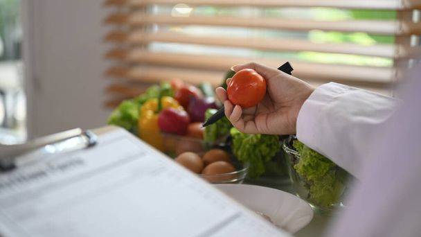 Ernährungswissenschaftlerin hält Tomate in der Hand und verschreibt Rezepte am Schreibtisch. Gesundheits- und Ernährungskonzept - Foto, Bild