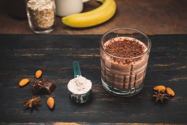 Milkshake smoothie, protein drink with grated chocolate in a glass on dark wooden board with bananas, protein powder in measuring spoon, star anise, almonds, cinnamon stick, chocolate pieces. - Photo, Image