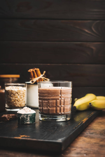Čokoládový koktejl, proteinový nápoj ve sklenici na tmavé dřevěné desce s banány, proteinový prášek v odměřovací lžičce a kousky čokolády. - Fotografie, Obrázek