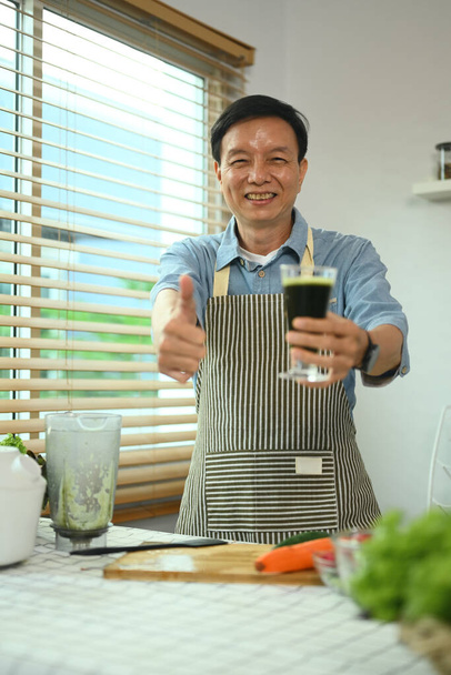 Fröhlicher Mann mittleren Alters hält Glas grünen Smoothie in der Hand und zeigt Daumen hoch - Foto, Bild