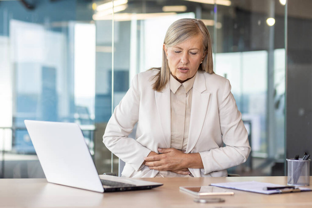 Sick overworked woman at workplace inside office has severe stomach pain, senior mature business woman holding hands on stomach massaging while sitting at desk with laptop. - Fotografie, Obrázek