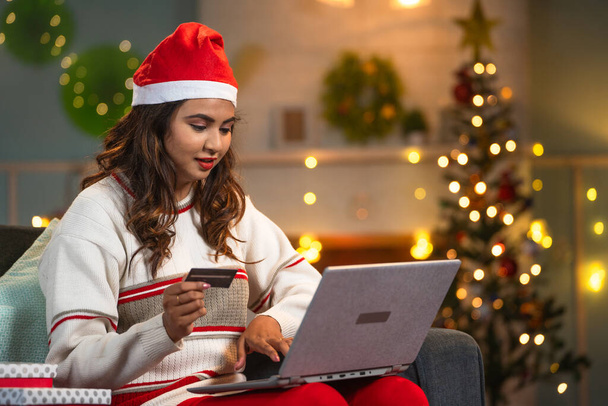 Счастливая женщина в шляпе Санта-Клауса делает покупки онлайн с помощью кредитной карты на ноутбуке во время празднования Рождества - концепция электронной коммерции, праздничные предложения или распродажи и банковские операции - Фото, изображение
