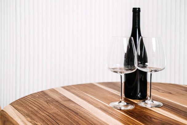Minimalistische wijnfles en glazen op houten tafel. Een eenvoudige maar elegante opstelling van een wijnfles en twee glazen op een gepolijste houten tafel tegen een gestreepte achtergrond - Foto, afbeelding