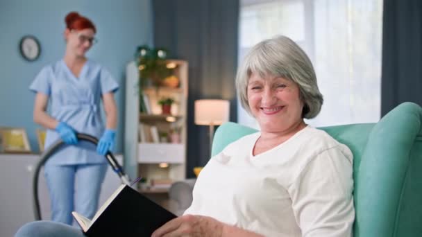 retrato de una anciana feliz con libro sonriendo y mirando el fondo de la cámara de la trabajadora social con una aspiradora en las manos - Metraje, vídeo