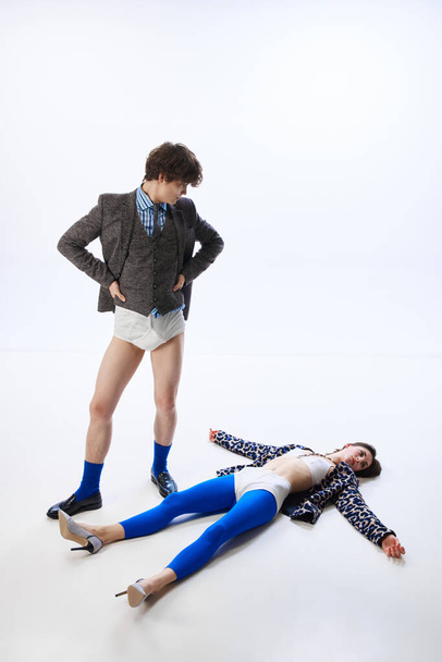 Young man in suit but without pants, in underwear, looks in bewilderment at girl who strangely and brightly dressed lying on floor. Concept of romance, bizarre love, Valentine Day, fashion, glamour - Photo, Image