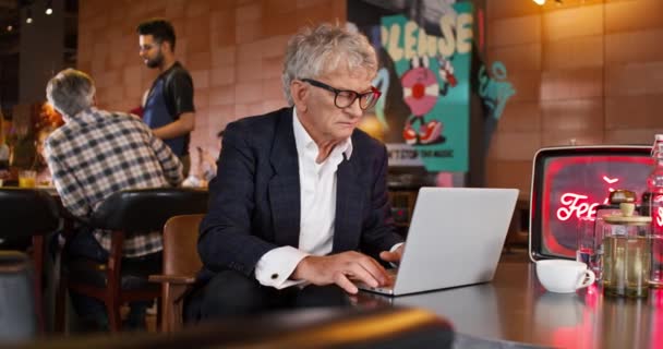 Starší obchodník s elegantním oblekem pracuje na svém přenosném notebooku, zatímco sedí v kavárně. V pozadí zákazníci komunikují nebo objednávají s číšníkem. Lidé odpočívající v restauraci. - Záběry, video