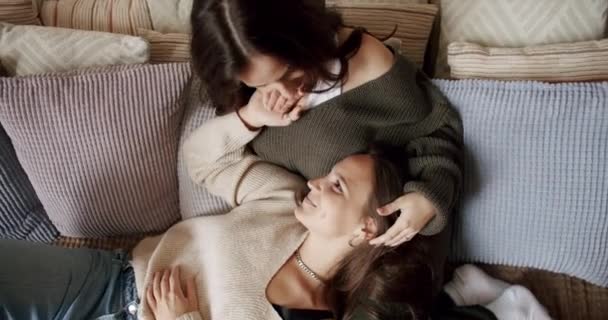 Vista superior de dos mujeres lesbianas tumbadas en un sofá con cojines, abrazándose y acariciándose mutuamente. Joven pareja femenina en relación romántica disfrutar de momentos de amor. Concepto de cercanía - Imágenes, Vídeo