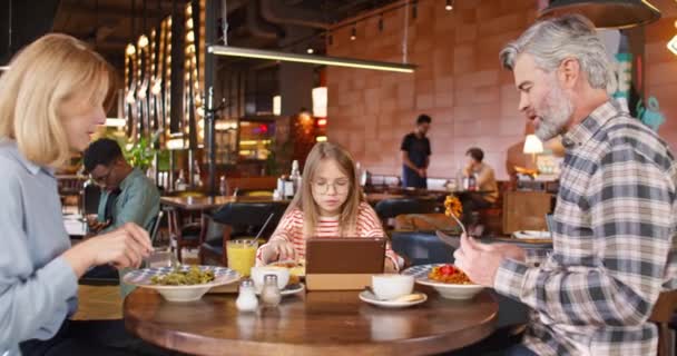 Zooming σε Καυκάσιος κορίτσι τρώει νόστιμο φαγητό, ενώ βλέποντας χαρτοκιβώτιο ή βίντεο στη συσκευή tablet. Αγαπημένη οικογένεια απολαμβάνοντας νόστιμο γεύμα στο καφέ. Γονείς επικοινωνούν κατά τη διάρκεια του δείπνου τους στο εστιατόριο. - Πλάνα, βίντεο